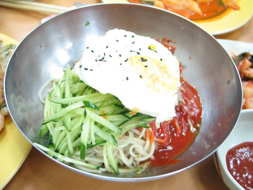 A bowl of spicy cold Korean noodles, bibim guksu, topped with a fried egg, cucumber, and sesame seeds