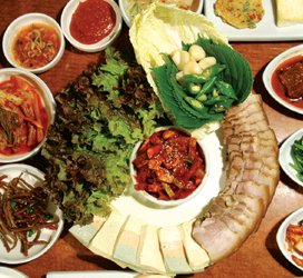 Sliced boiled pork belly fanned on a plate next to napa cabbage leaves and fresh kimchi for wrapping