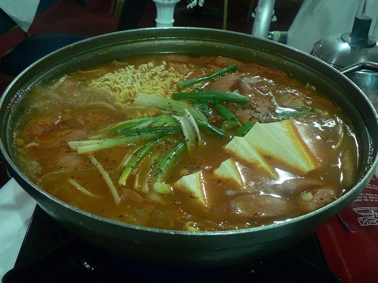 Budae jjigae simmering in a shallow pot with visible sausage and kimchi