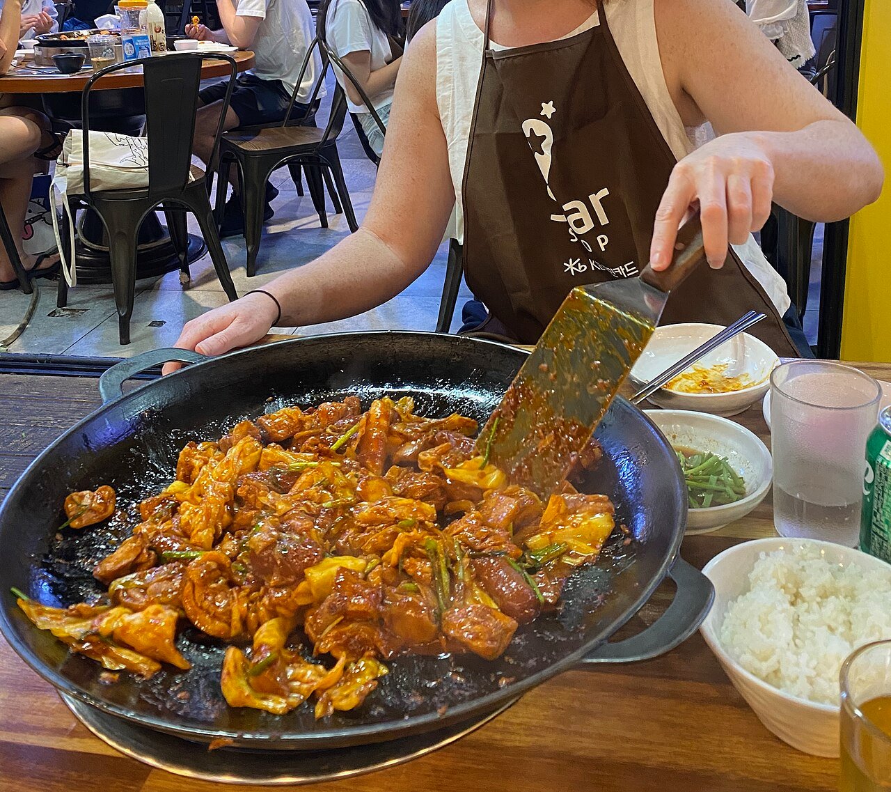 Chicken and vegetables being stirred with metal spatulas in a large pan on a tabletop burner