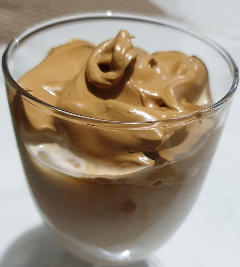A glass of iced milk topped with a thick layer of pale brown whipped dalgona coffee foam
