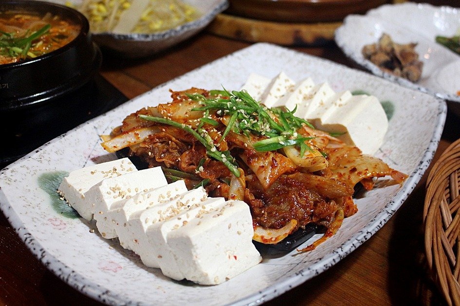 Close view of stir-fried kimchi with pork belly next to warm tofu slices