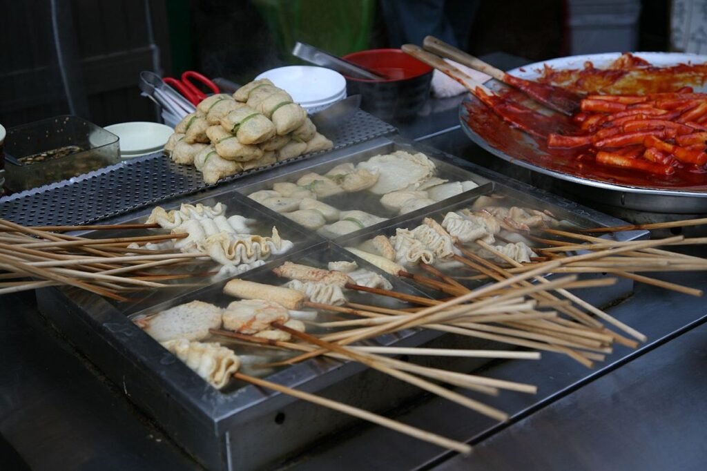 A street food stall at Pupyeong Market in Busan with fish cake skewers standing in a steaming broth tray beside a pan of tteokbokki