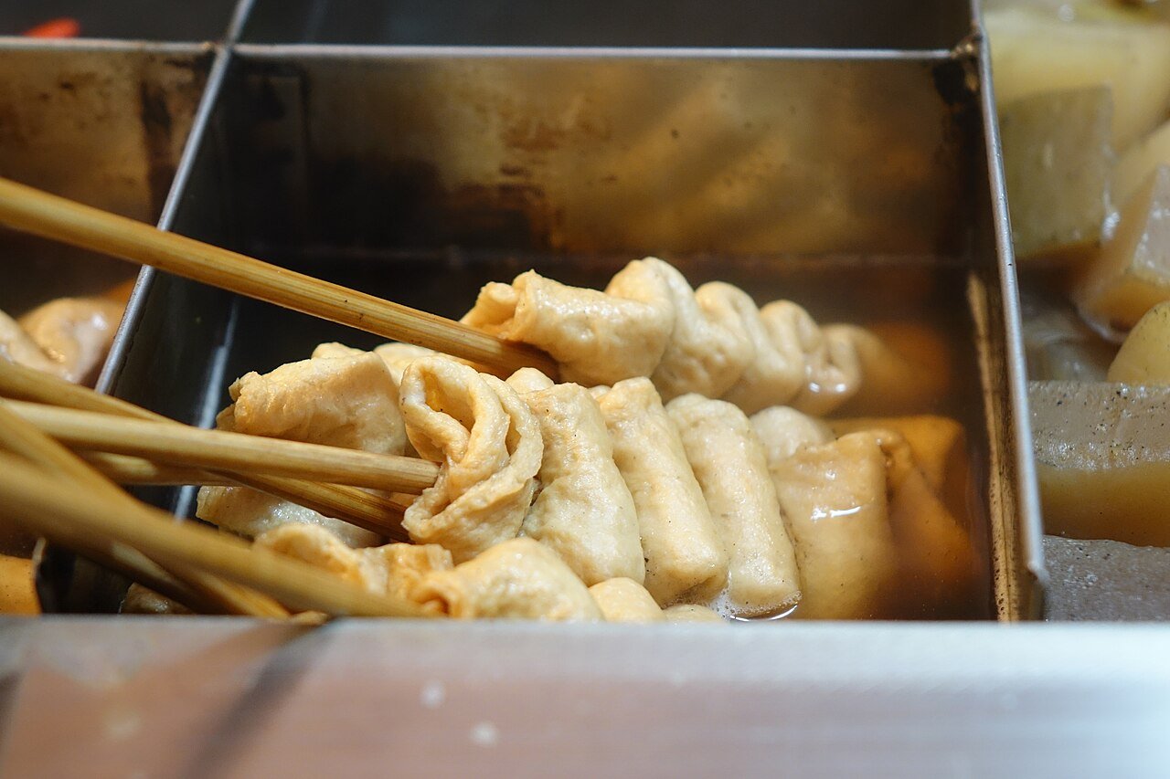 Folded fish cake skewers simmering in a metal broth tray at a street stall, with tongs lifting one skewer out