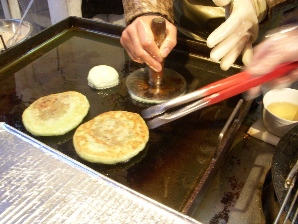 A round stainless steel press with a wooden handle flattening filled hotteok dough on an oiled griddle.