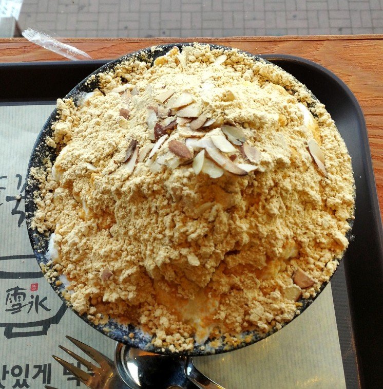 A bowl of shaved ice dessert topped with cubes of injeolmi rice cake and a dusting of soybean powder