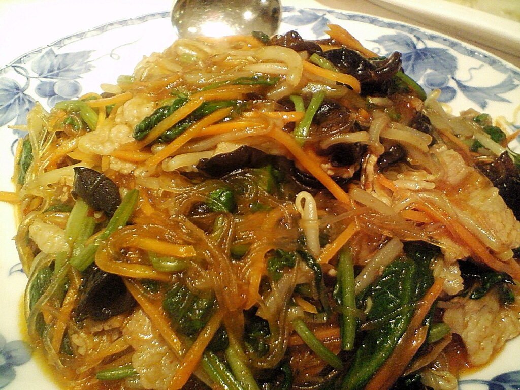 A plate of japchae, glass noodles tossed with beef, spinach, carrot and mushroom in a shallow serving dish