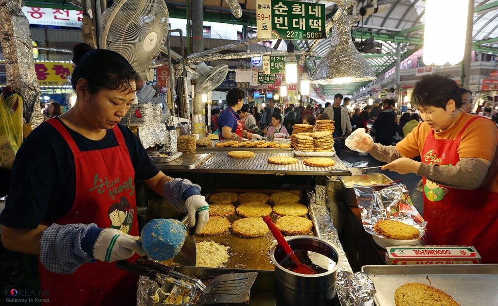 Two vendors in red aprons frying mung bean pancakes on a griddle at a covered market stall in Seoul