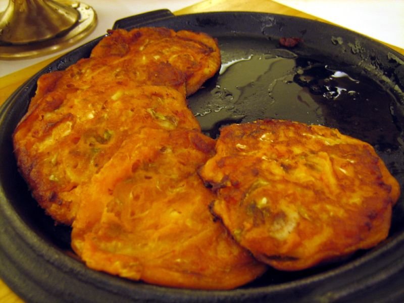 A golden-brown kimchi jeon pancake studded with red kimchi, cut into wedges on a plate