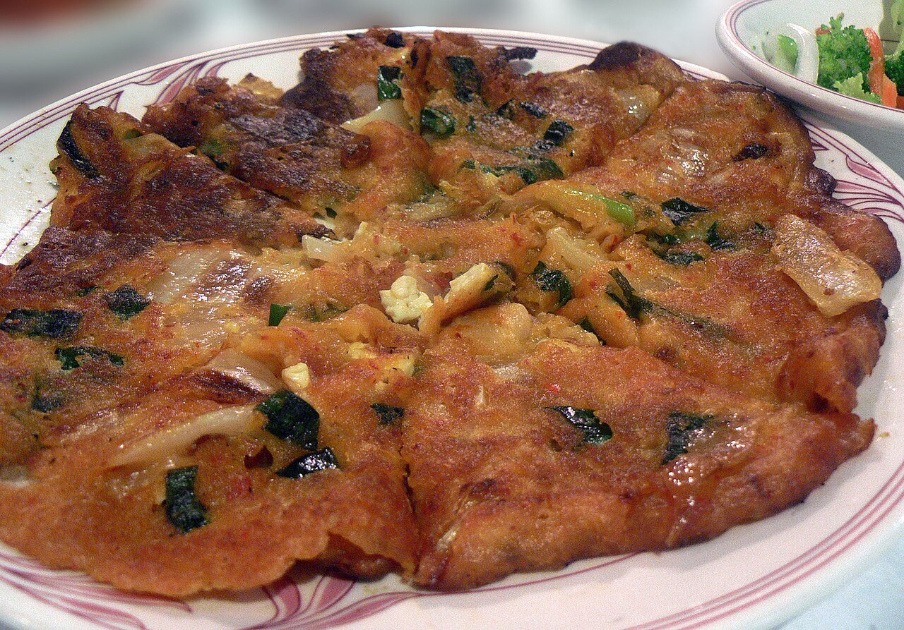 A whole kimchi pancake in a pan, red kimchi pieces visible through the batter