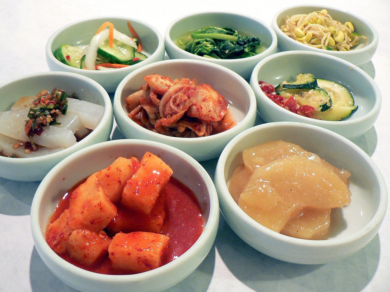 Six small side dishes of Korean banchan arranged in two rows on a table, including cucumber salad and kimchi