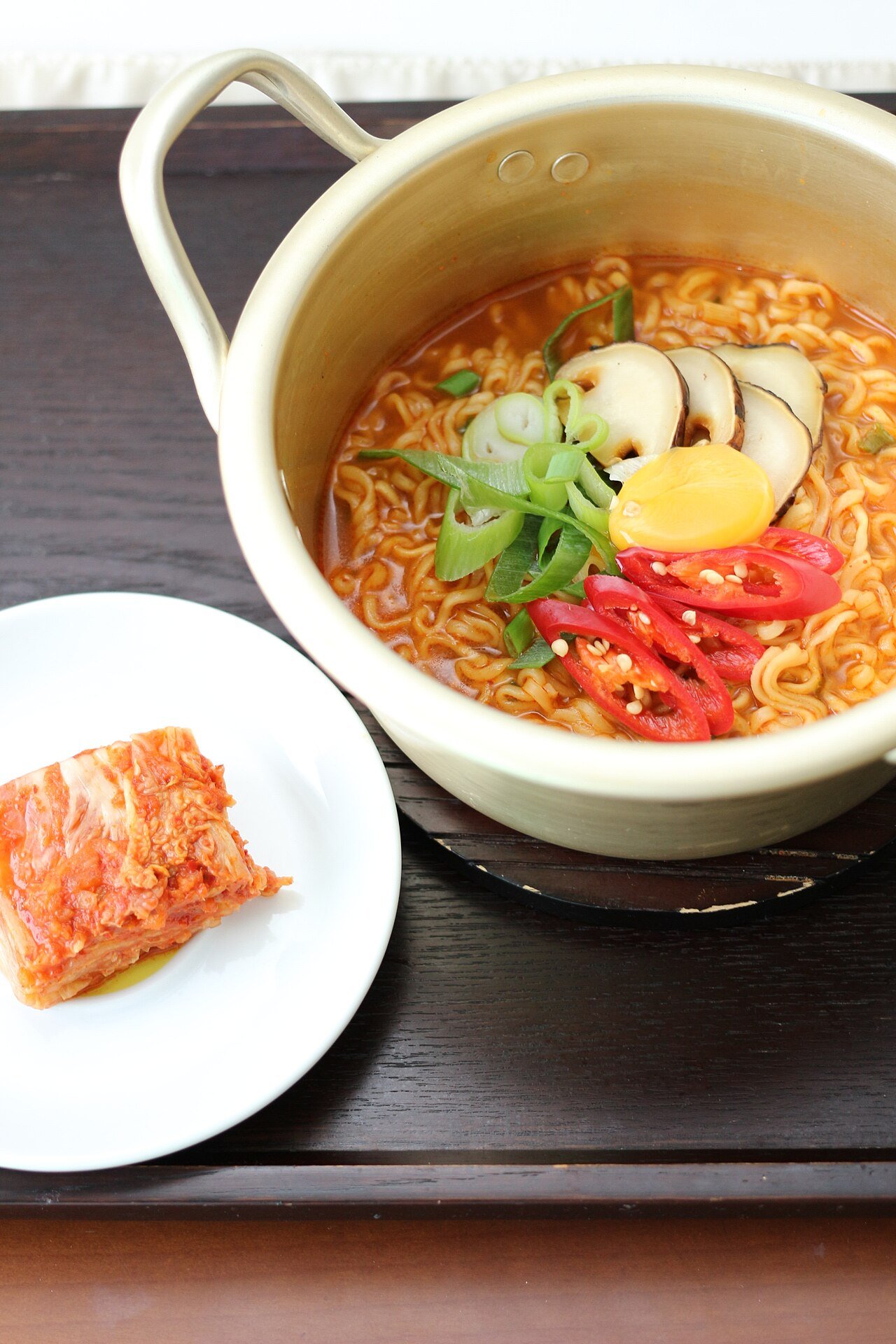 A pot of Korean ramyeon served with a small dish of napa cabbage kimchi beside it