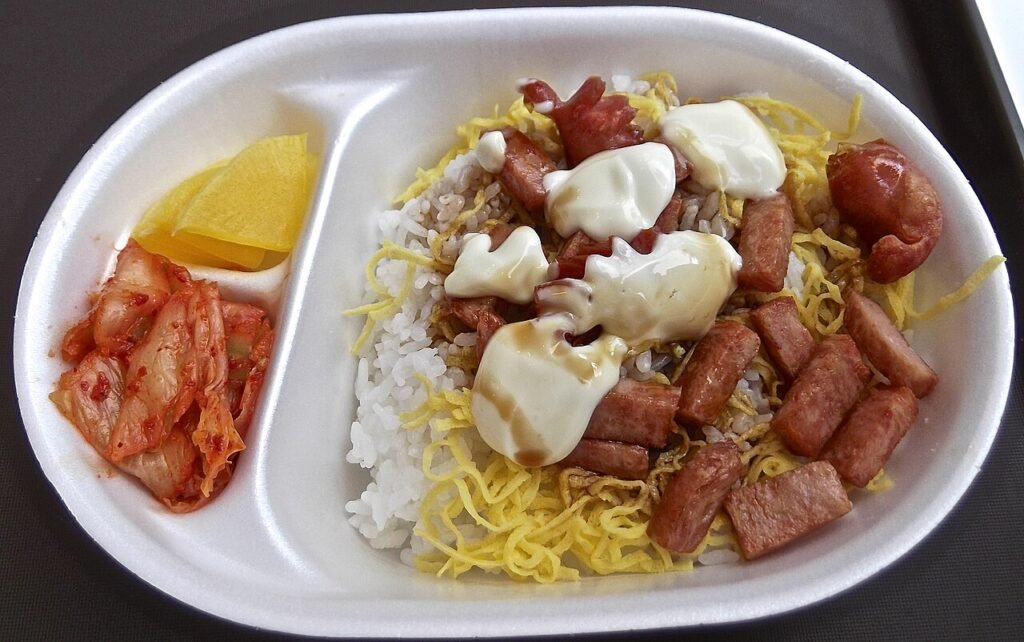 A compartmentalized Korean lunch box with rice, ham, and side dishes