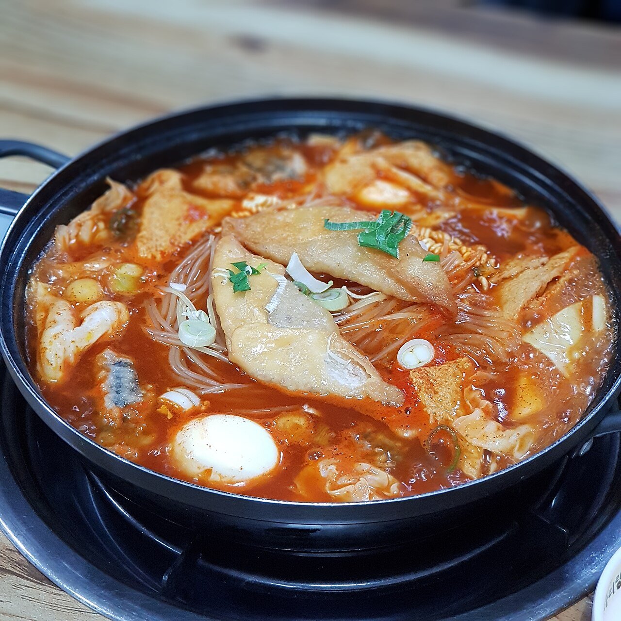 A wide shallow pan of rice cakes, fish cake sheets, cabbage and noodles simmering in red sauce at a table