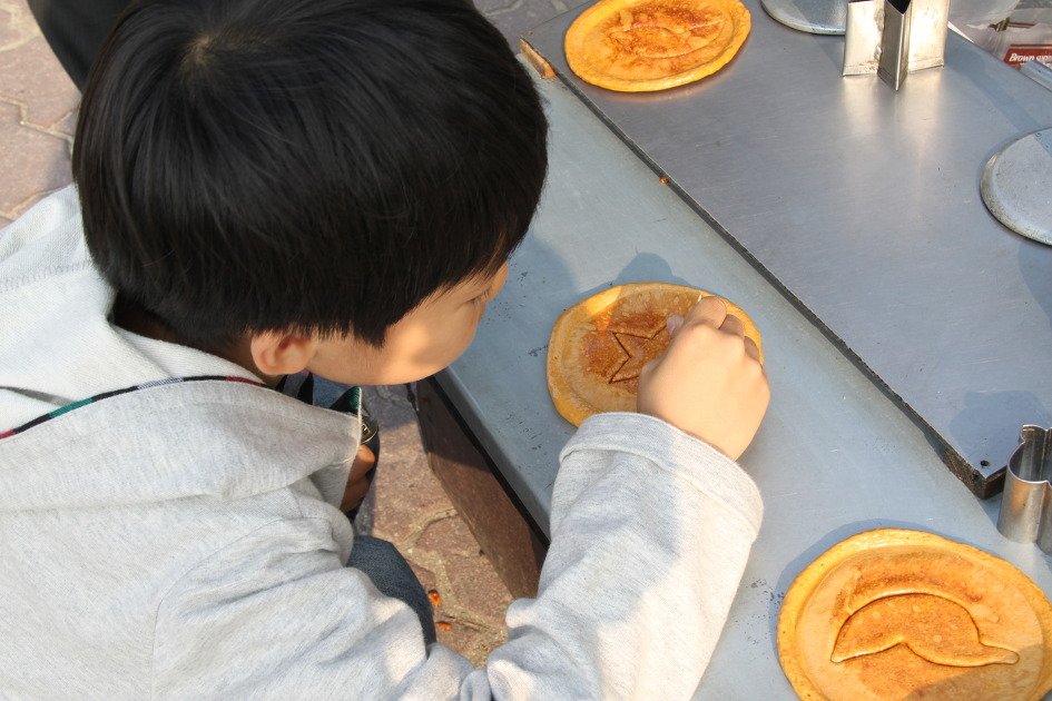 A child using a needle to trace a star shape stamped into a flat honeycomb-colored dalgona candy on a metal stall counter