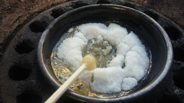 White sugar melting into clear syrup in a small metal ladle set over a charcoal burner, stirred with a wooden stick