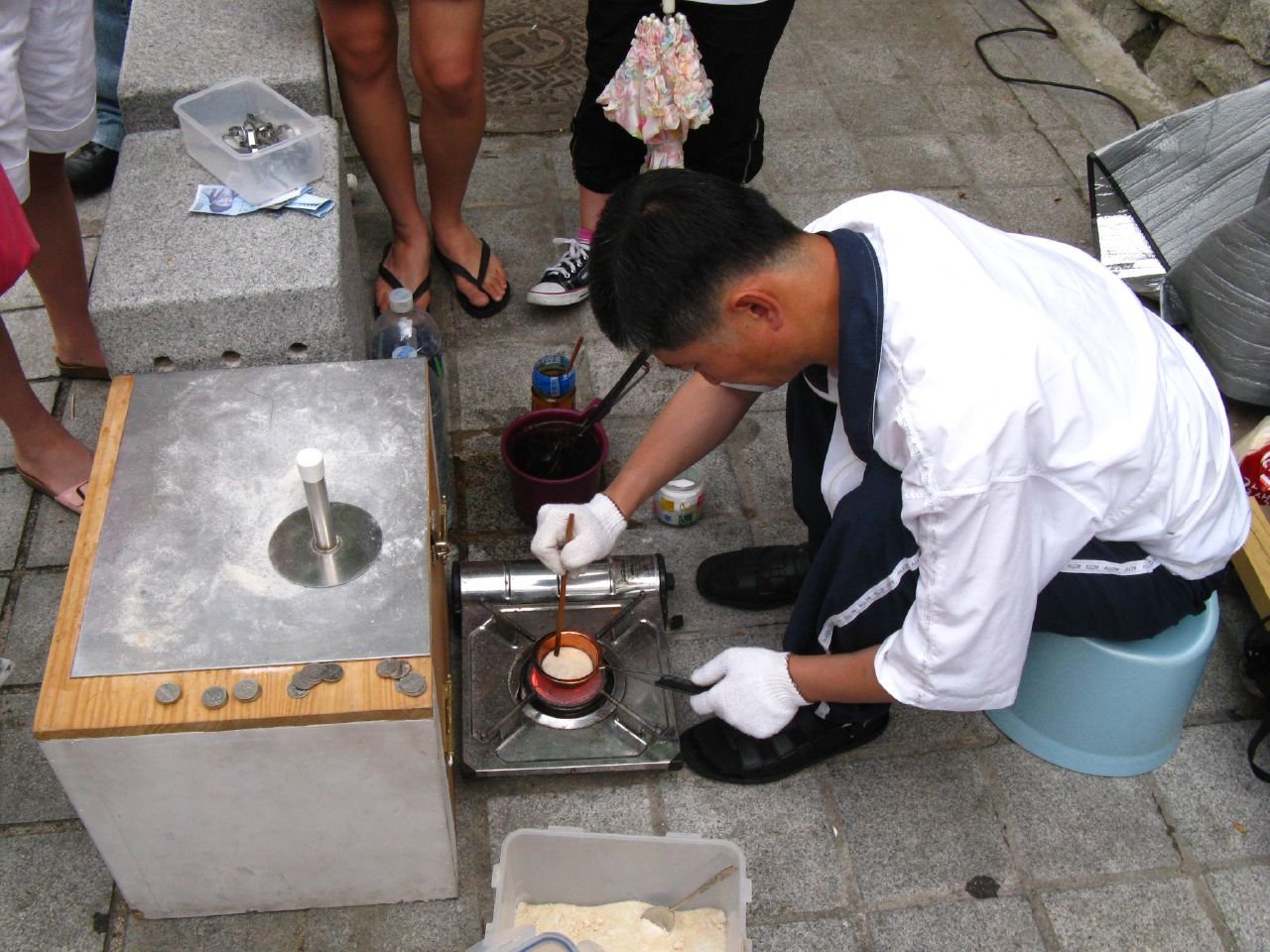 A street vendor in white gloves melting sugar in a ladle over a portable gas burner beside a metal pressing box and coins
