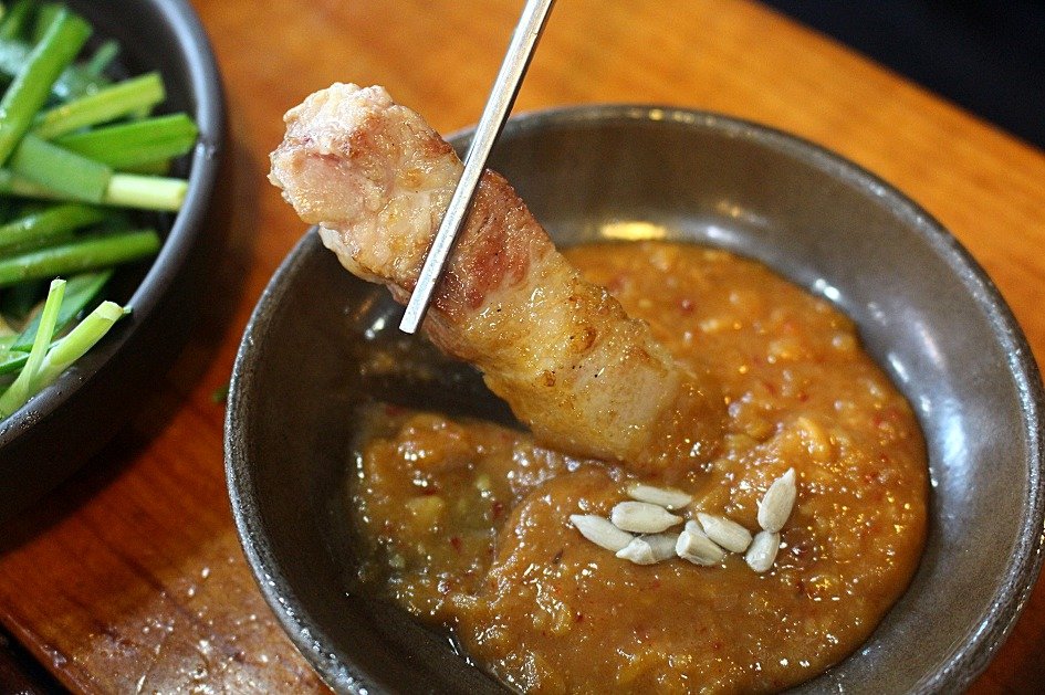A piece of grilled pork belly being dipped into a small bowl of ssamjang