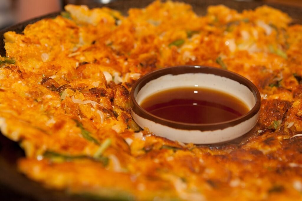 A crisp Korean seafood and scallion pancake with a small bowl of thin brown dipping sauce set in the middle