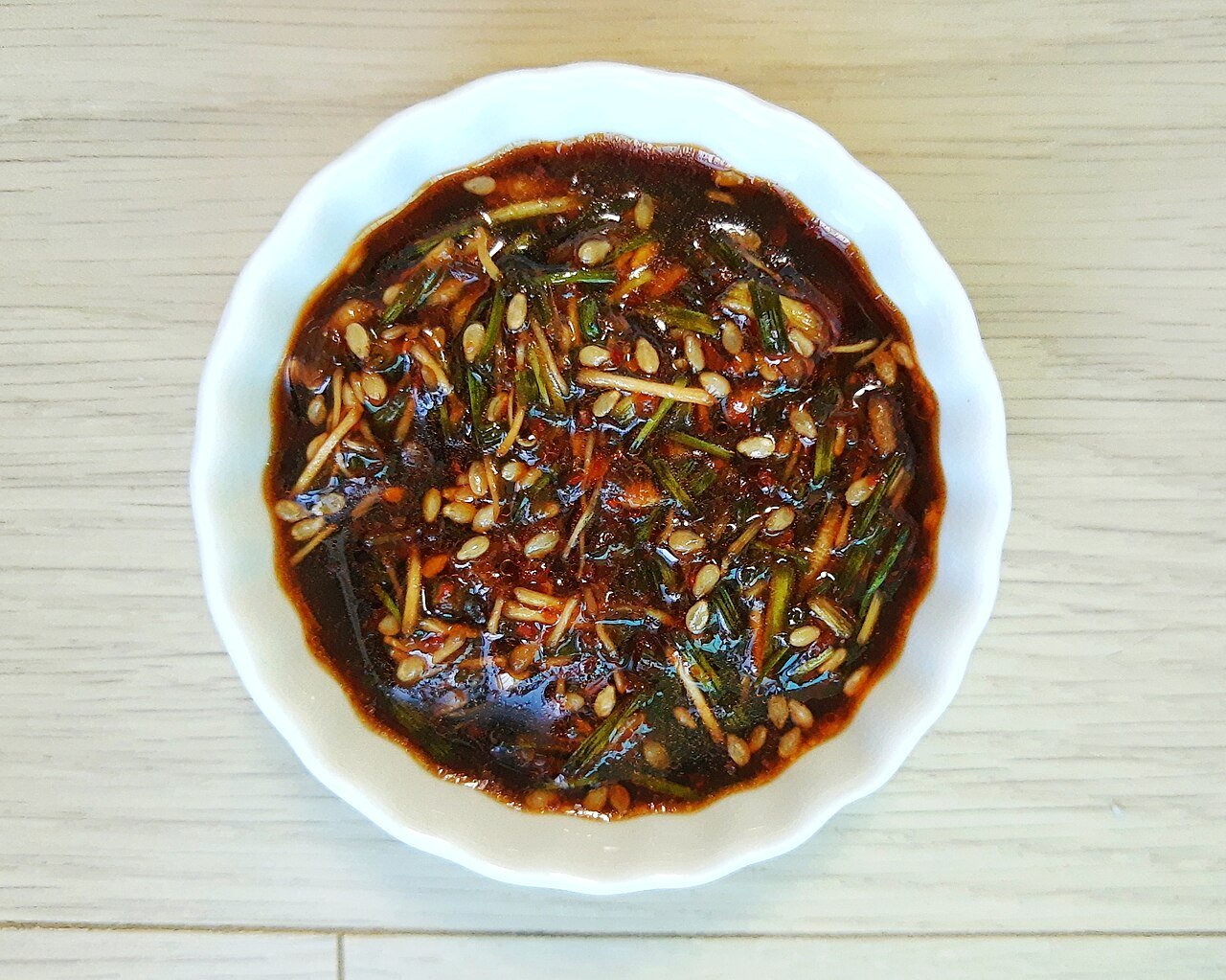 A white dish of dark soy-based Korean seasoning sauce with chili flakes, chopped greens and sesame seeds