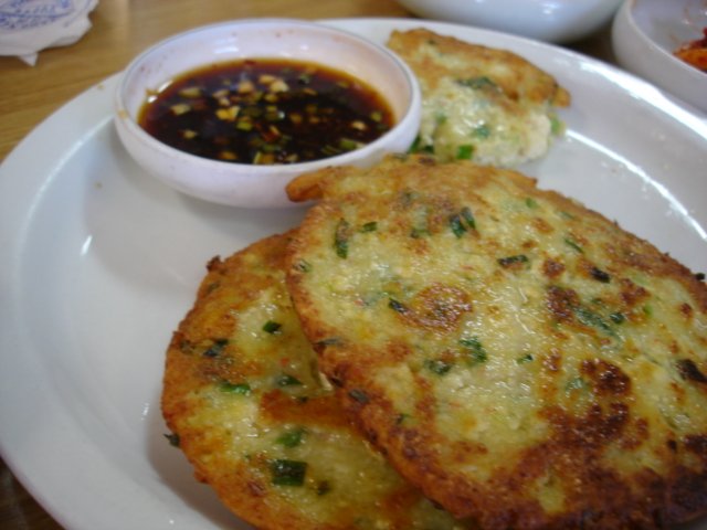 A plate of pan-fried Korean tofu pancakes beside a small white dish of soy dipping sauce flecked with garlic and scallion