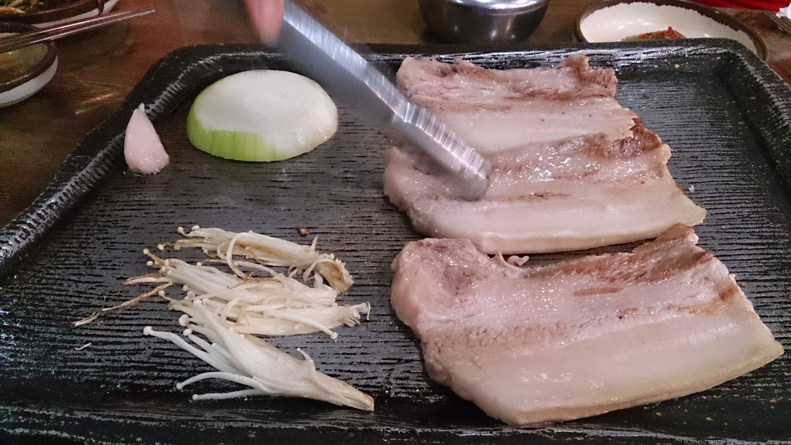 Tongs turning thick slices of pork belly on a black grill plate next to a halved onion, a garlic clove and enoki mushrooms