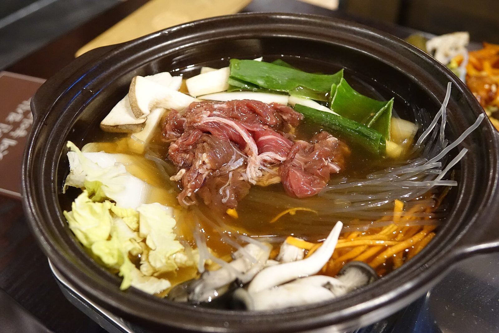 Black clay pot of bulgogi hot pot with marinated beef, glass noodles, mushrooms, cabbage and scallions in broth