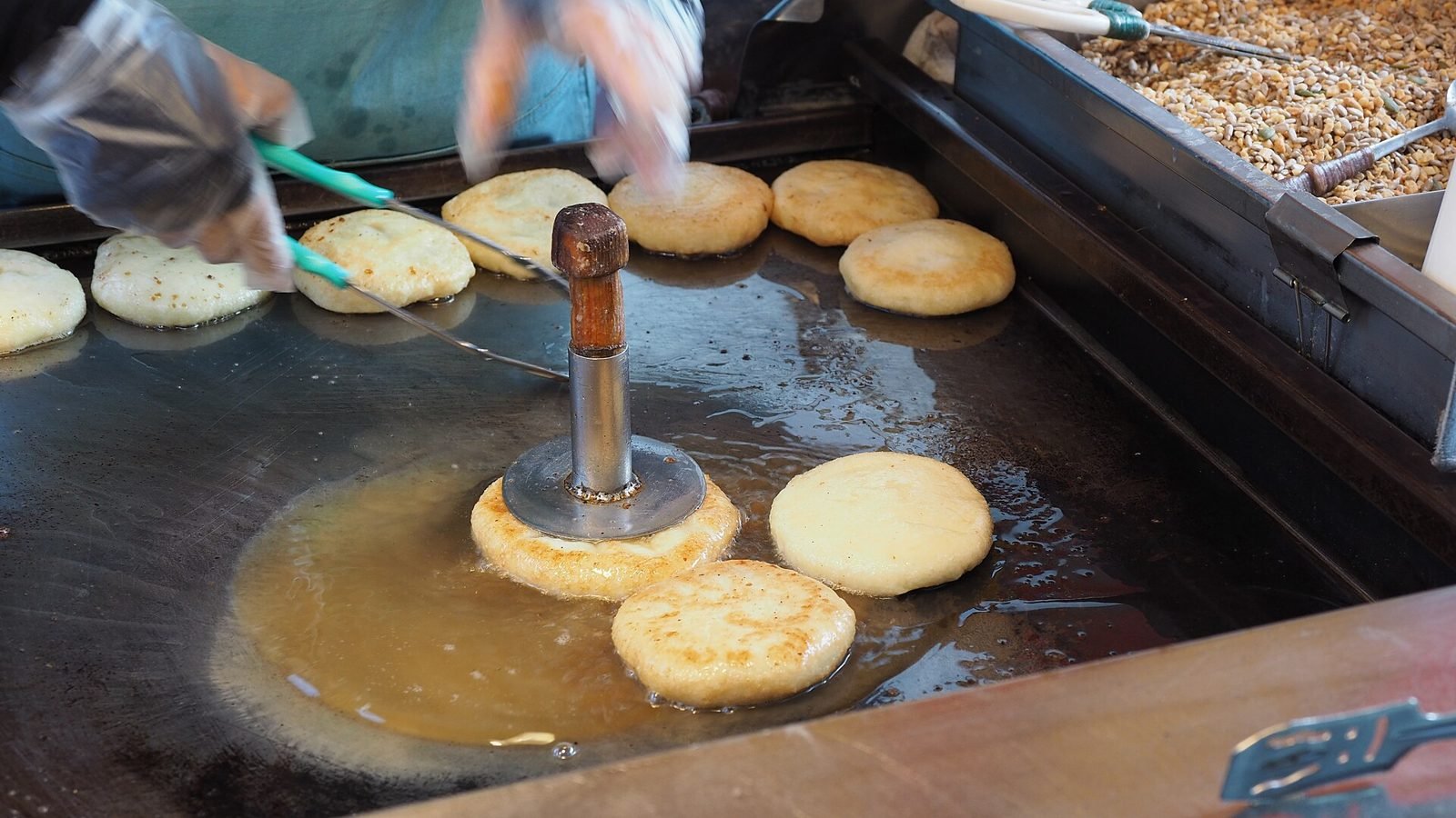 Street vendor pressing hotteok dough balls flat with a metal disc on a griddle