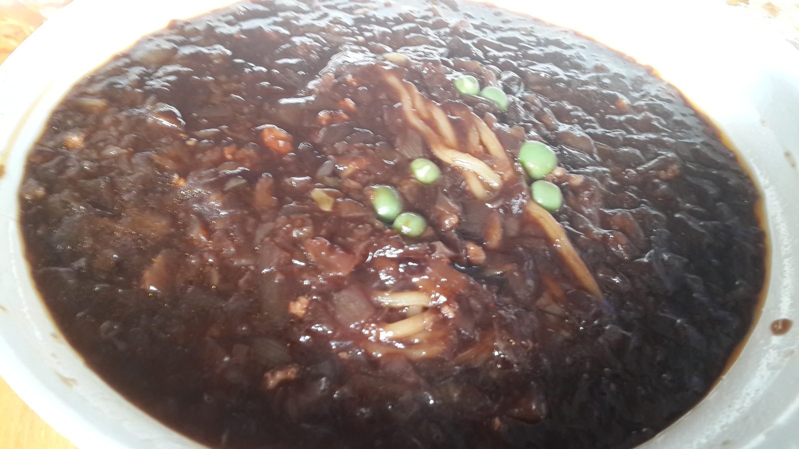 Close view of a bowl of jjajangmyeon covered in thick glossy black sauce with diced onion and sliced green topping