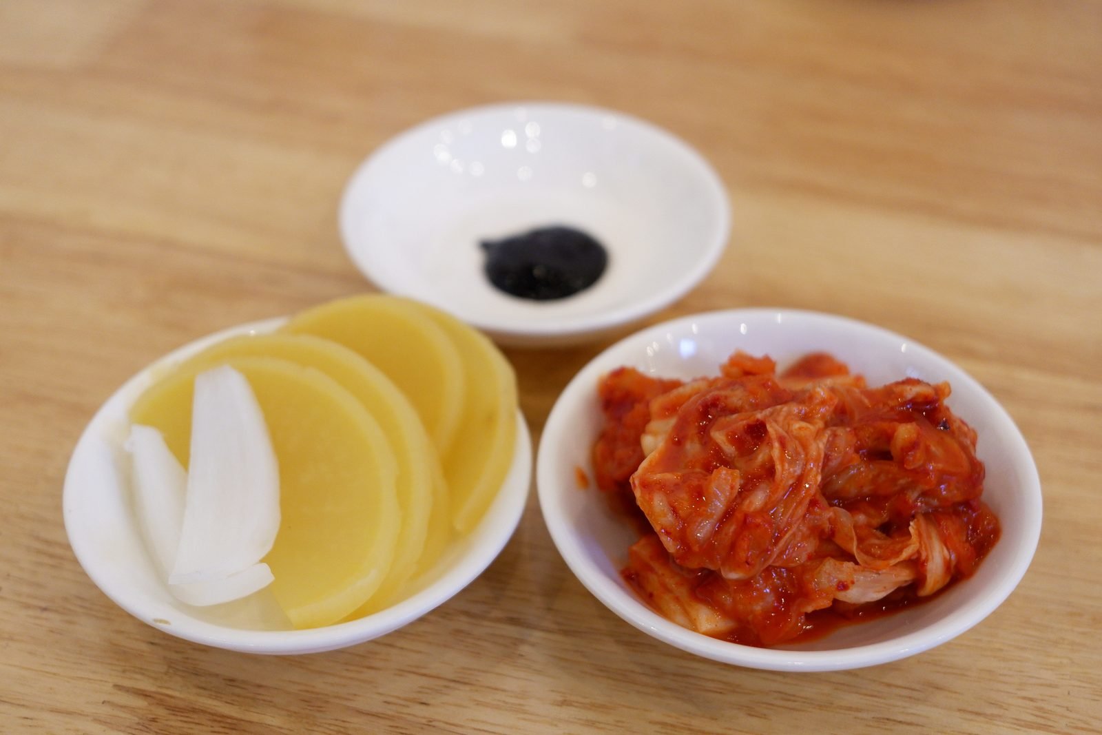 Three small white dishes holding black chunjang paste, yellow pickled radish with raw onion, and red kimchi on a wooden table