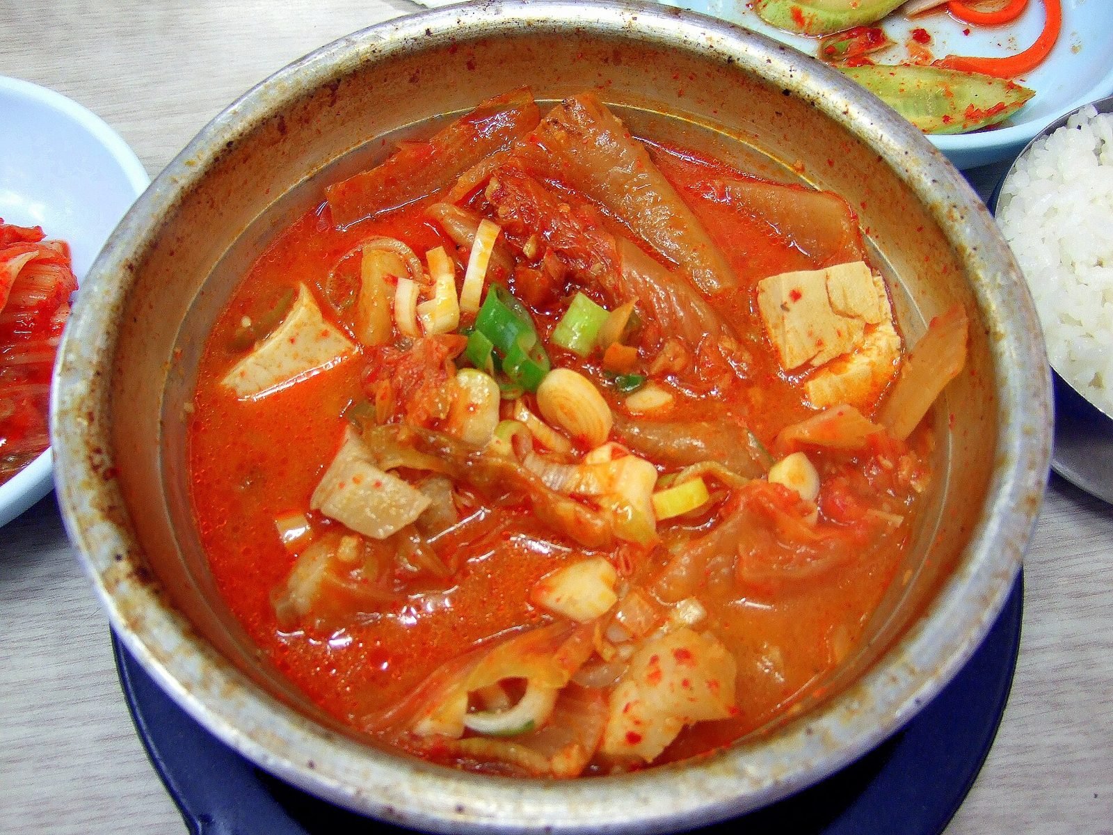 Metal bowl of red kimchi stew with tofu and scallions beside rice