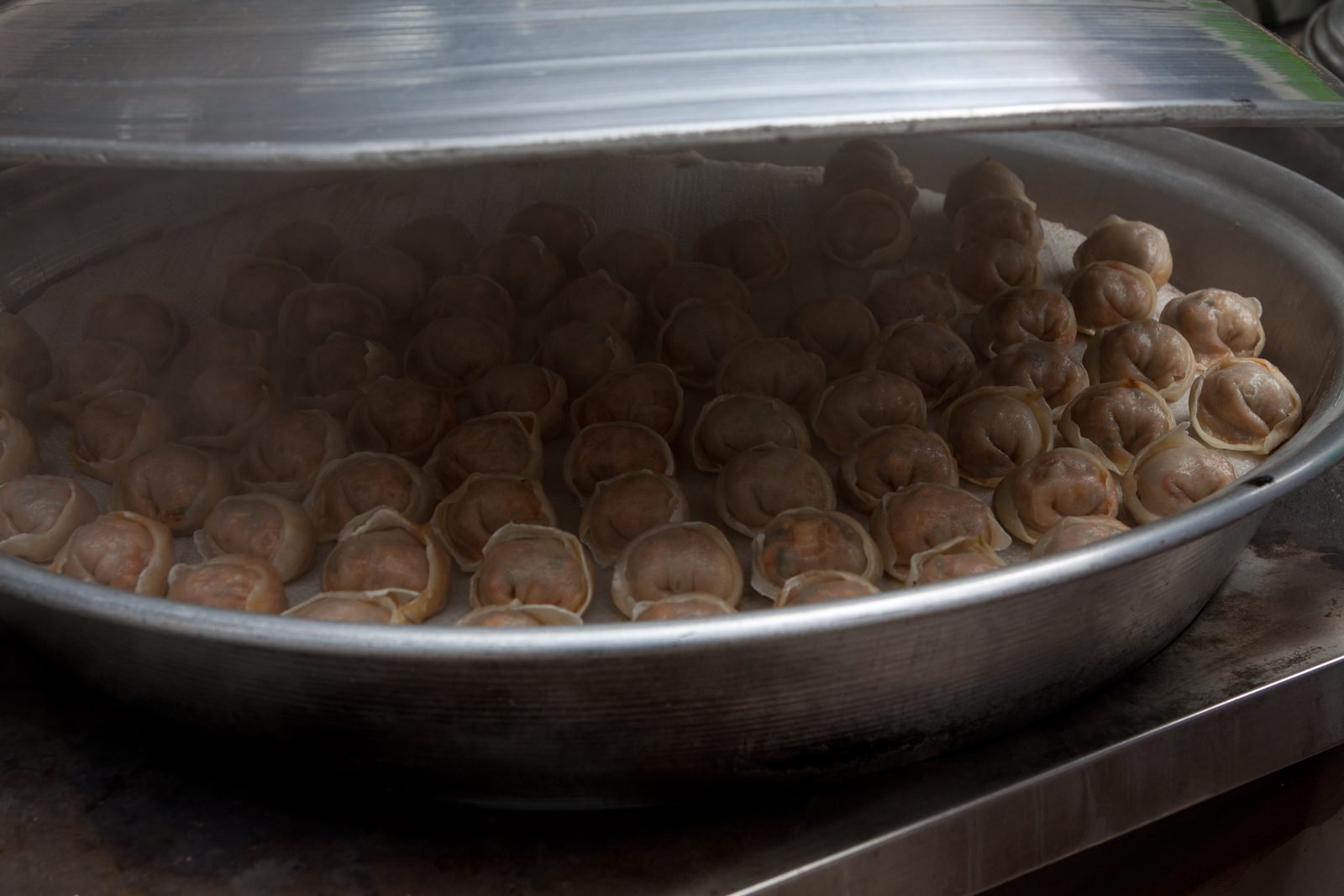 Rows of steamed Korean dumplings packed in a large metal steamer tray with its lid lifted