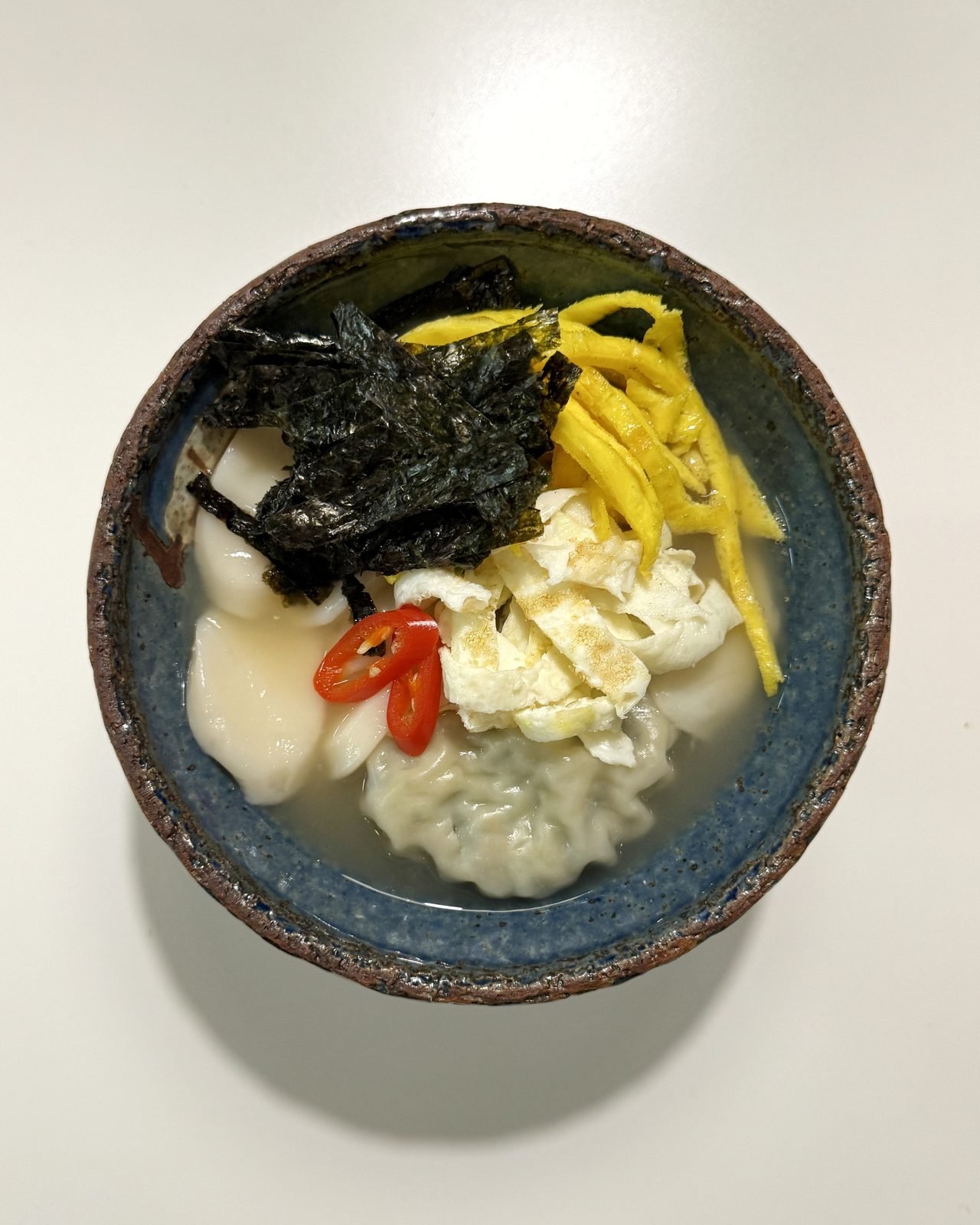 Blue ceramic bowl of tteok-mandu-guk with rice cake slices, a dumpling, seaweed strips, egg ribbons and red chili