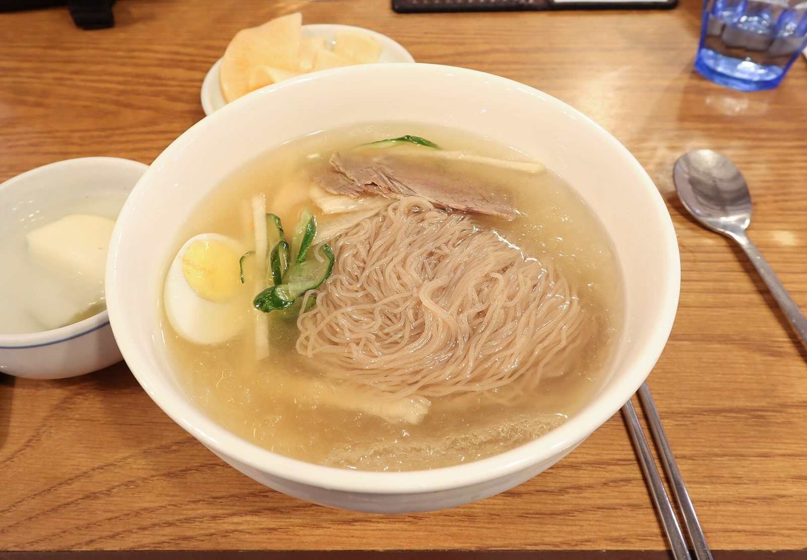 White bowl of Pyongyang-style naengmyeon in clear broth with thin noodles, sliced beef, cucumber and half a boiled egg
