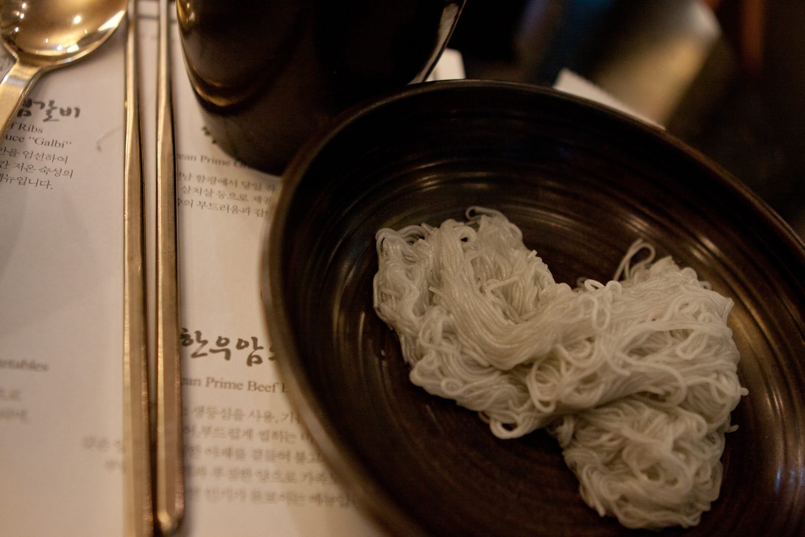 A loose nest of thin pale gray naengmyeon noodles in a dark round bowl beside a menu and chopsticks