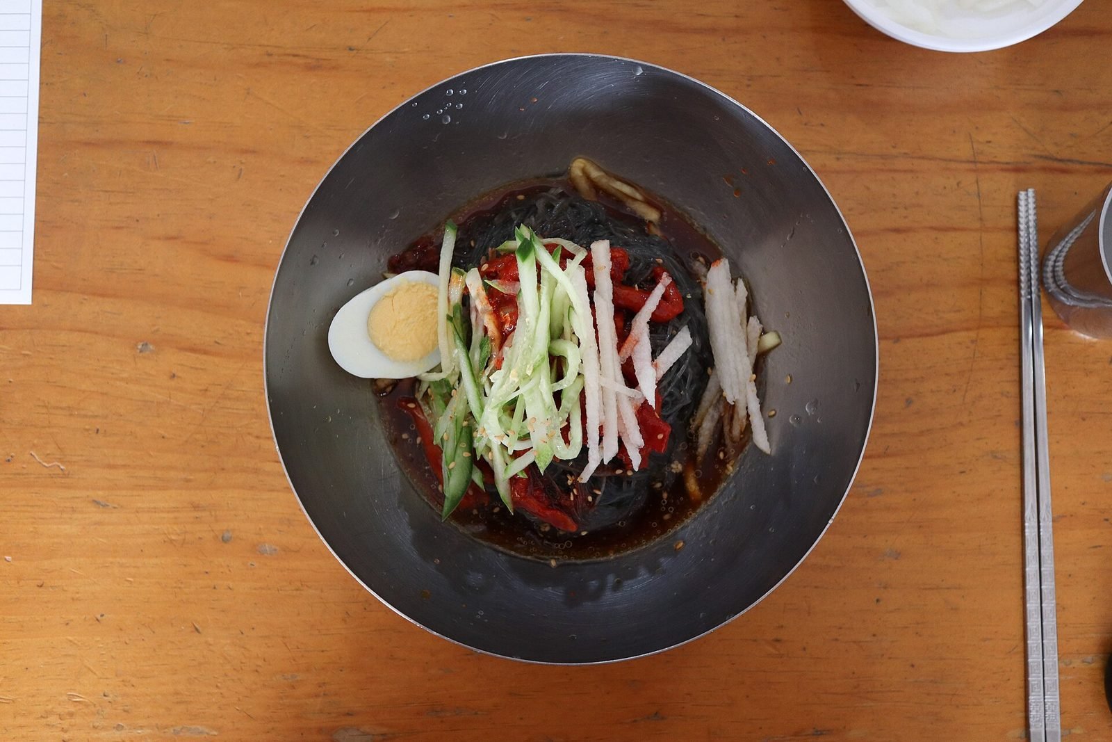 Steel bowl of dark naengmyeon noodles in red chili sauce topped with cucumber, pear strips and half a boiled egg