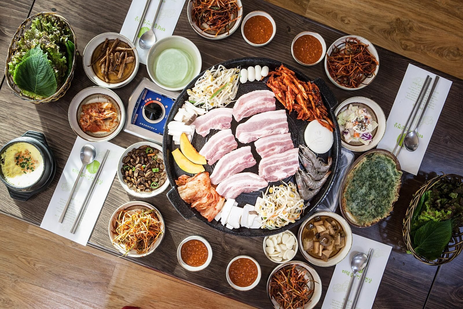 Overhead view of a table with pork belly on a grill surrounded by small side dishes and lettuce
