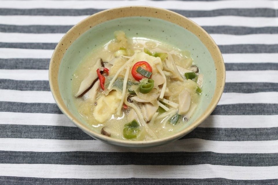 Pale creamy sujebi with mushrooms and sliced green chili in a celadon bowl on a striped cloth