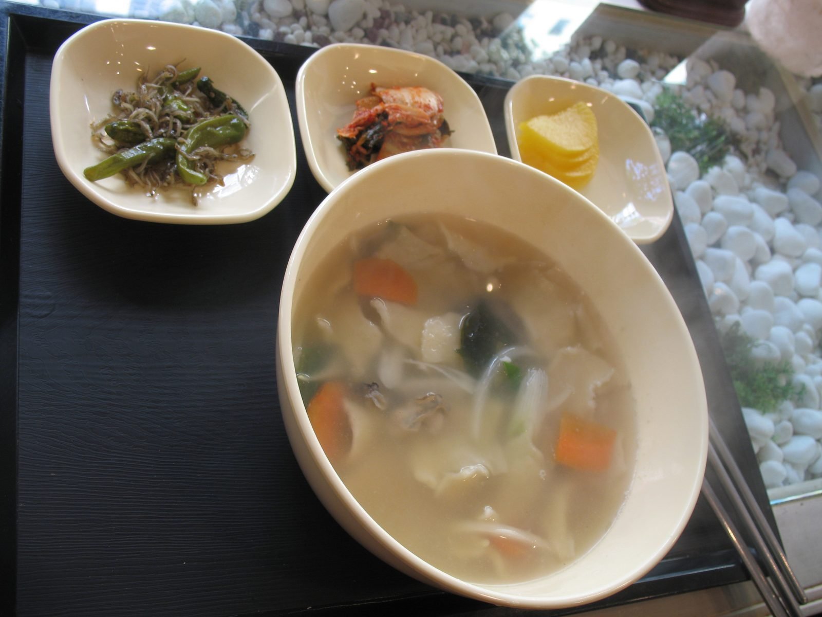 Bowl of sujebi, a clear broth with torn dough pieces, potato and carrot, beside small side dishes on a tray